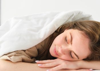 Portrait of happy young woman sleeping in her bed, enjoying a nap in hotel suite, lying under blanket.