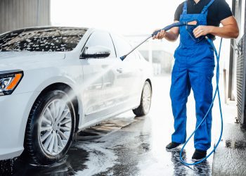 Professional washer in blue uniform washing luxury car with water gun on an open air car wash. Close up photo.