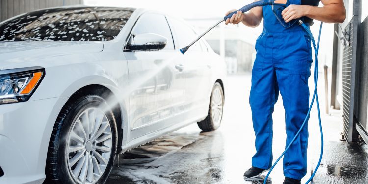 Professional washer in blue uniform washing luxury car with water gun on an open air car wash. Close up photo.