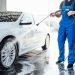 Professional washer in blue uniform washing luxury car with water gun on an open air car wash. Close up photo.
