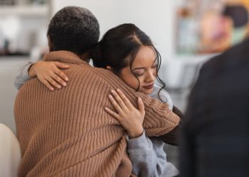 A young mixed-race woman hugs a mature adult black woman. They are sitting next to each other in a medical clinic. The two women are attending a group therapy session. They are showing support and kindness.