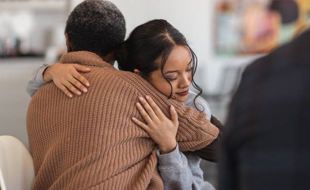 A young mixed-race woman hugs a mature adult black woman. They are sitting next to each other in a medical clinic. The two women are attending a group therapy session. They are showing support and kindness.