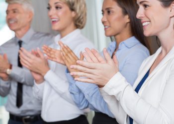 Audience of four business people looking at somebody, smiling, standing and applauding. Side view.