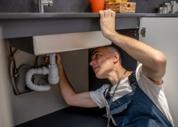 Plumber, repair. Experienced attentive middle-aged man carefully examining bottom of kitchen sink touching pipe with his hand diagnosing problem