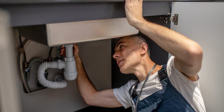 Plumber, repair. Experienced attentive middle-aged man carefully examining bottom of kitchen sink touching pipe with his hand diagnosing problem