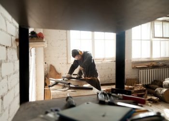 Trained young workman grinding metal parts of construction on table in workshop interior with tools equipment. Manual worker wearing protective clothes working with angle grinder in manufactory