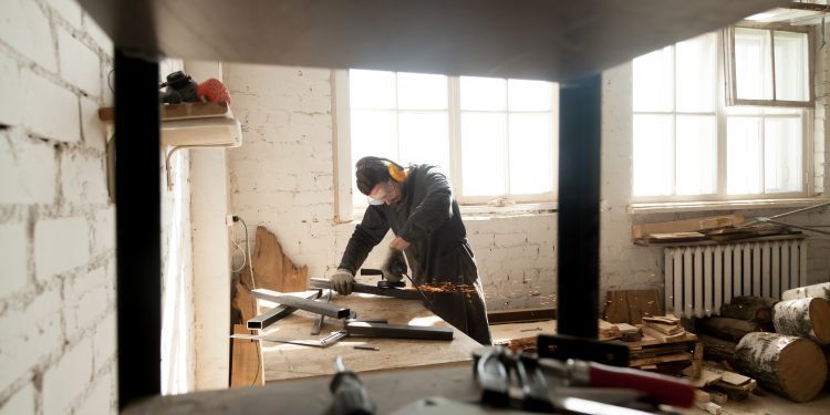 Trained young workman grinding metal parts of construction on table in workshop interior with tools equipment. Manual worker wearing protective clothes working with angle grinder in manufactory
