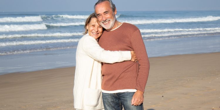 Beautiful senior couple standing at seashore on sunny day, smiling and looking at camera. Happy blonde woman hugging gray-haired bearded man, showing affection. Love, holiday, retirement concept