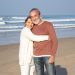 Beautiful senior couple standing at seashore on sunny day, smiling and looking at camera. Happy blonde woman hugging gray-haired bearded man, showing affection. Love, holiday, retirement concept