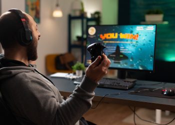 Player winning video games with controller and headset in front of monitor. Man using joystick and headphones, playing online games on computer. Person celebrating game win for leisure.