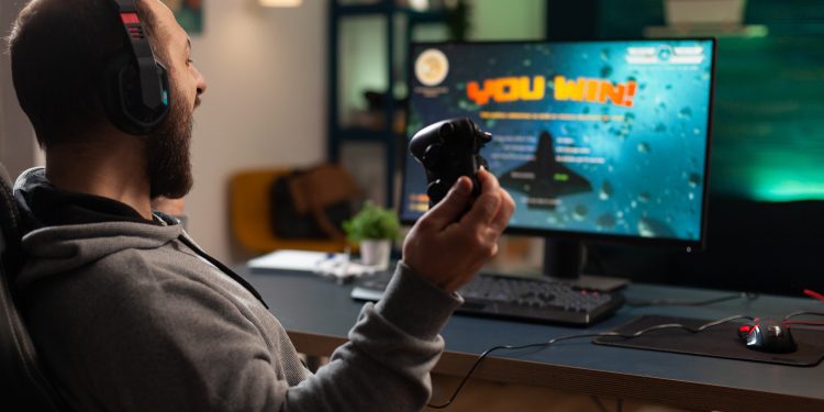 Player winning video games with controller and headset in front of monitor. Man using joystick and headphones, playing online games on computer. Person celebrating game win for leisure.