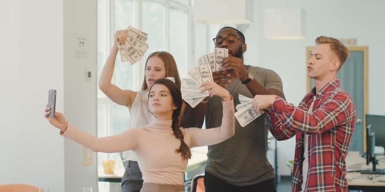 Young people making fun with money at coworking space. Happy business people with money taking selfie at workplace. Cheerful team dancing with dollar banknote looking at camera in office.