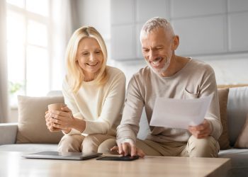 A joyful elderly couple engaged in reviewing financial documents while relaxing at home with cups of coffee