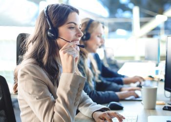 Female customer support operator with headset and smiling, with collegues at background
