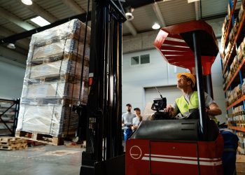 Forklift operator loading cargo while working in a warehouse. His colleagues are in the background.