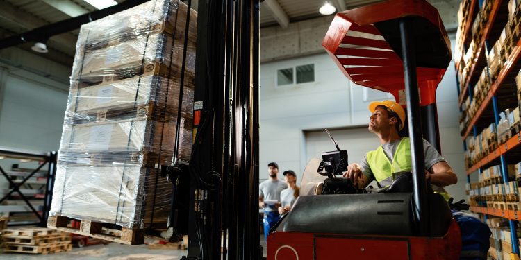 Forklift operator loading cargo while working in a warehouse. His colleagues are in the background.