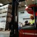 Forklift operator loading cargo while working in a warehouse. His colleagues are in the background.