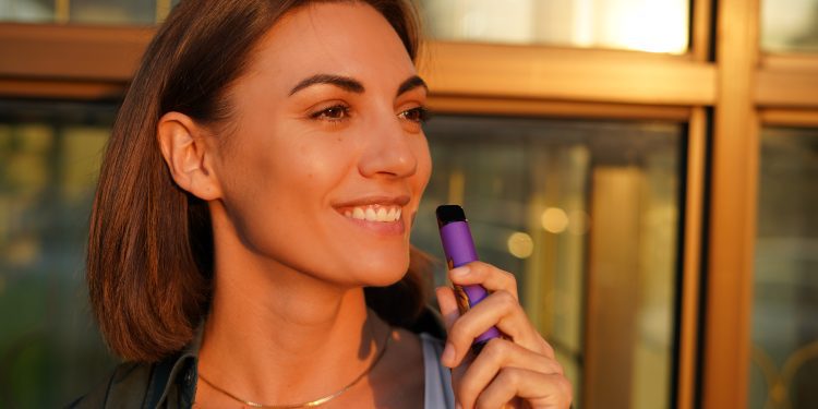 Woman with cup of coffee outdoor at city street at sunset with electronic cigarette catching sun rays enjoying smoking