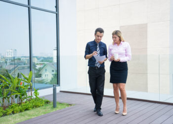 Two confident business colleagues walking on office terrace. Mid adult business man and woman discussing document outdoors on coffee break. Working during break concept