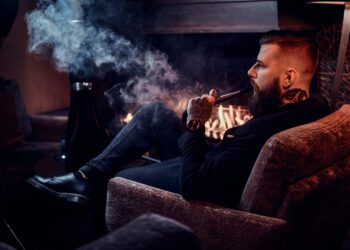 Bearded relaxed man is sitting on the armchair and smoking hookah near fireplace.