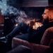 Bearded relaxed man is sitting on the armchair and smoking hookah near fireplace.