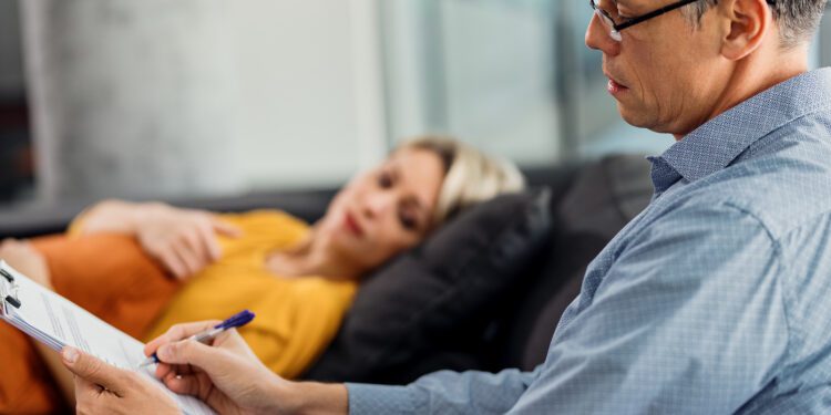 Mental health professional taking notes while having therapy session with a woman at his office.