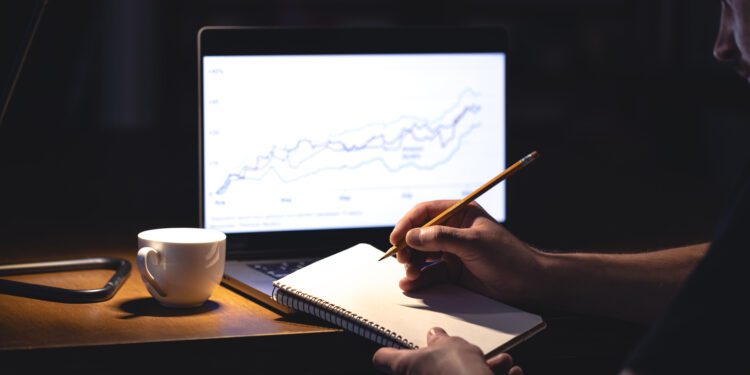 A man sits in front of a laptop with a chart and notepad at night. Business man working with computer and analysis graph stock market trading with stock chart data.