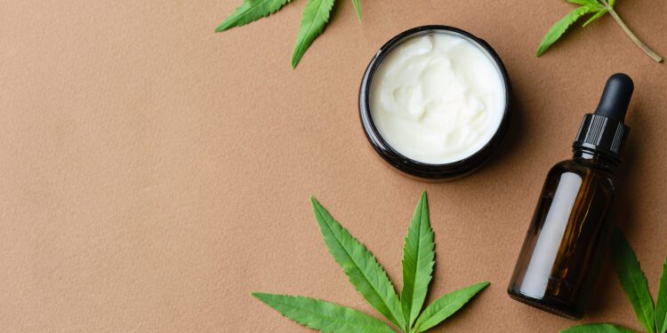 Top view of Cannabis cosmetic oil cream in jar bottle and a green plant leaf Natural cosmetic on brown background. Flat lay, copy space