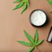 Top view of Cannabis cosmetic oil cream in jar bottle and a green plant leaf Natural cosmetic on brown background. Flat lay, copy space