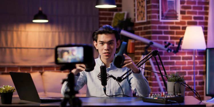 Asian influencer filming headphones review on podcast episode, recording recommendation video on camera. Live broadcasting talk show with wireless headset product on social media.