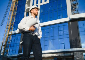 Successful engineer or architect, crane and building construction at backgrpound. Joyous businessman wearing helmet