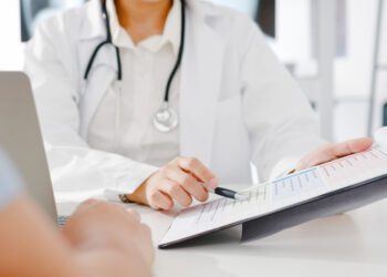 Young Asia female doctor in white medical uniform using clipboard is delivering great news talk discuss results or symptoms with male patient sitting at desk in health clinic or hospital office.