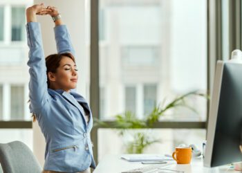 Young smiling businesswoman stretching with her eyes closed while working on a computer in the office.