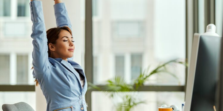 Young smiling businesswoman stretching with her eyes closed while working on a computer in the office.
