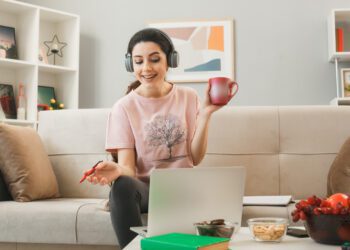 smiling young girl holding cup of tea with pen wearing headphones used laptop sitting on sofa behind coffee table in living room