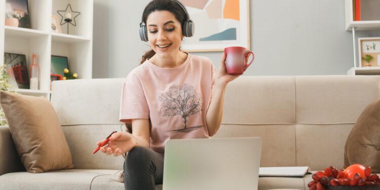 smiling young girl holding cup of tea with pen wearing headphones used laptop sitting on sofa behind coffee table in living room