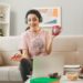 smiling young girl holding cup of tea with pen wearing headphones used laptop sitting on sofa behind coffee table in living room